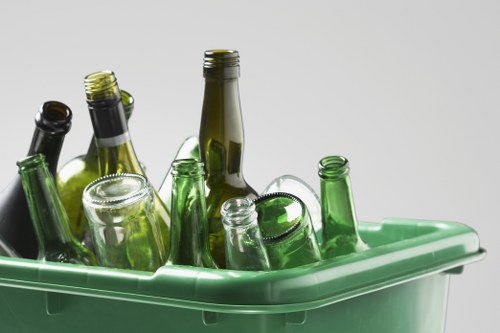 A collection of empty glass bottles of varying sizes and shapes, predominantly in shades of green and brown, with some clear glass bottles, all placed inside a light green plastic bin. The bottles are upright, with some leaning against each other, and their open necks and mouths are visible. The bin appears to be situated on a neutral, light-colored surface with a plain, light gray background, indicating an indoor or controlled environment. The image has a focus on the bottles' textures and transparency, highlighting their smooth glass surfaces. This scene is relevant to rubbish removal, specifically recycling or disposal of glass waste, as represented by the company Commercial Waste Coney Hall, which offers such services in the local area near Coney Hall.