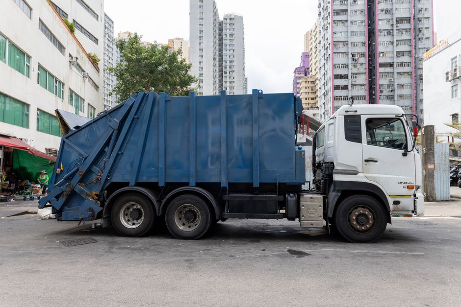 A flatbed truck equipped with a compact hydraulic lift system is parked on a paved street in front of a modern, multi-storey residential building. The truck's cabin is orange with black detailing, and