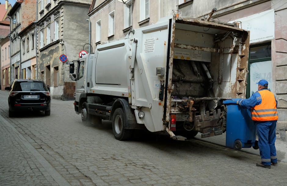 Two large black garbage bags that appear to be made of plastic or similar material are tied at the top and placed on the pavement near a black metal fence. The bags are filled with waste, causing them