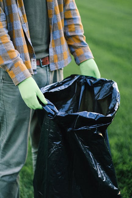A large, weathered green waste skip filled with a mixture of cardboard boxes, paper bags, and various debris, situated outdoors on a paved area. The cardboard items include open-backed boxes of differ